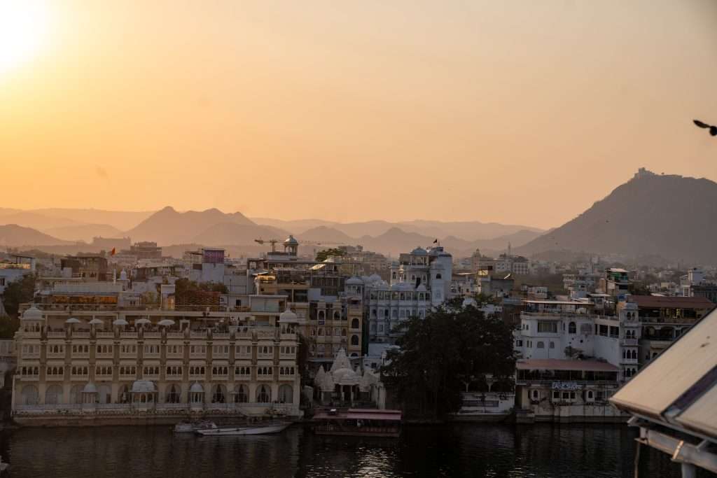 A serene view of a city at sunset, featuring a blend of historic and traditional buildings near a calm body of water. Hills and mountains can be seen in the background, fading into the distance under a warm, golden sky. Birds are faintly visible in the sky.