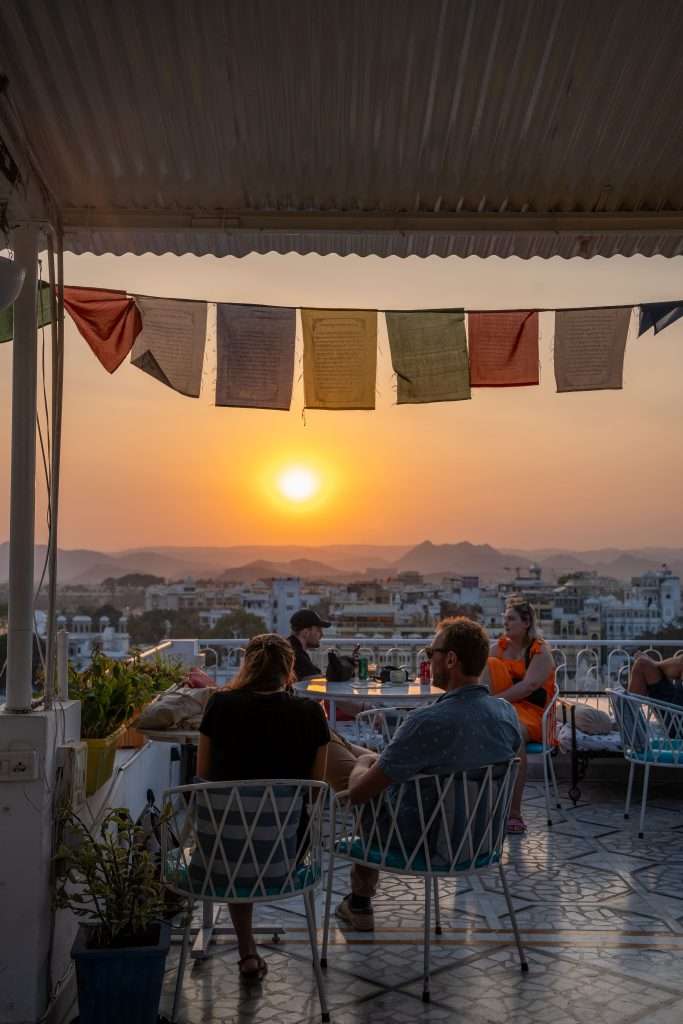 People sit on a rooftop terrace at sunset, with colorful prayer flags overhead. The view includes a distant cityscape and mountains, bathed in the warm glow of the setting sun. Tables and chairs are arranged across the tiled floor of the terrace - Dreamyard Hostel Udaipur