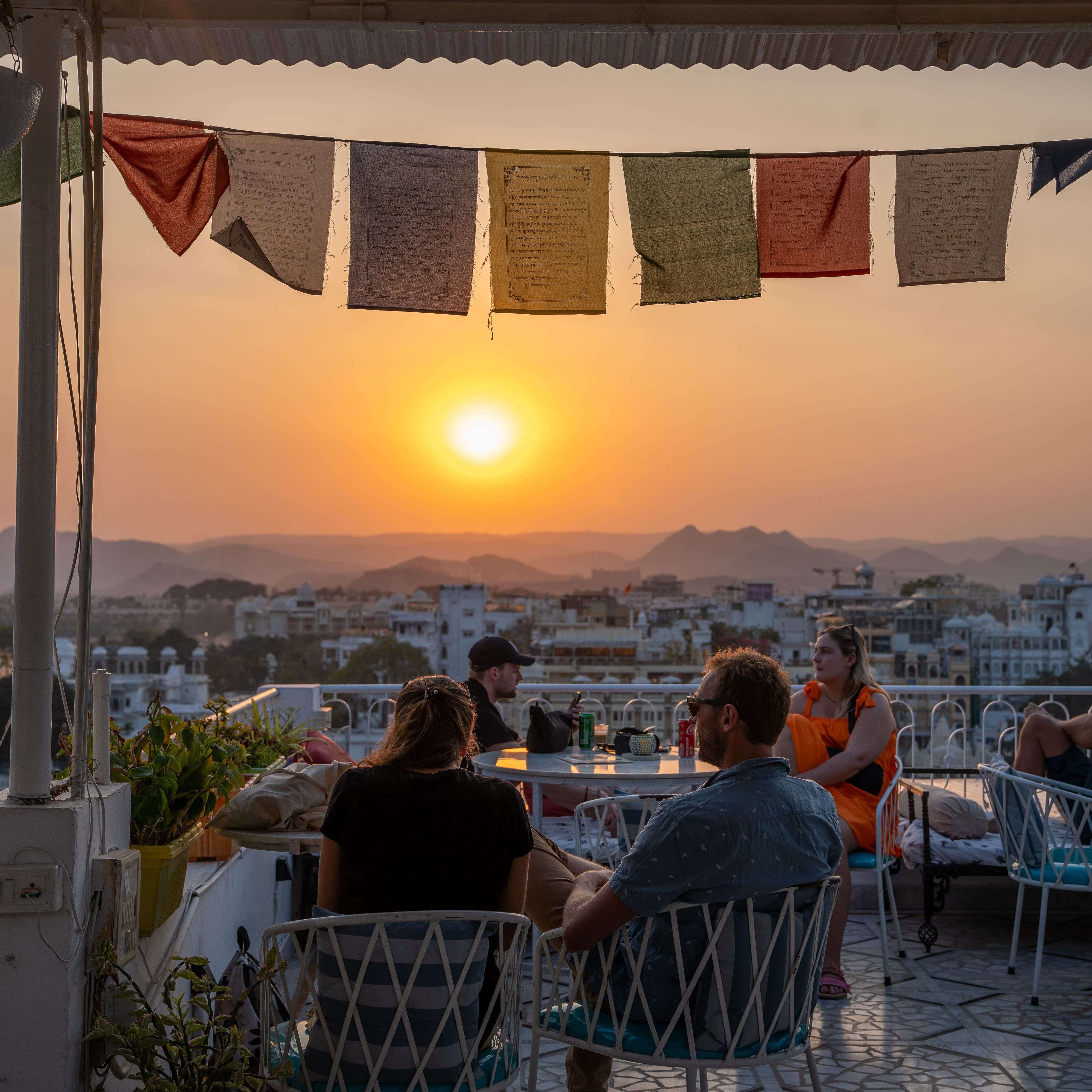 People sit on a rooftop terrace at sunset, with colorful prayer flags overhead. The view includes a distant cityscape and mountains, bathed in the warm glow of the setting sun. Tables and chairs are arranged across the tiled floor of the terrace - Dreamyard Hostel Udaipur