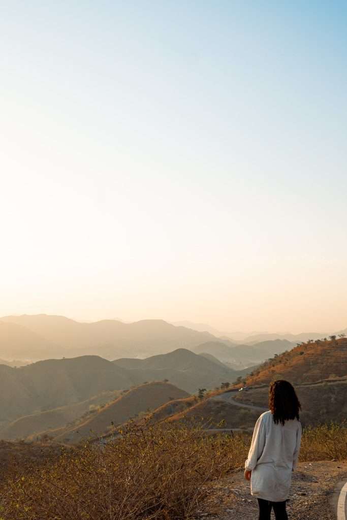 A person with long hair stands on a hill overlooking a valley with rolling hills and mountains under a clear sky at sunset, wearing a light-colored sweater and dark pants. Sparse vegetation is visible in the foreground, making one ponder, "Is Udaipur worth visiting for such serene beauty?