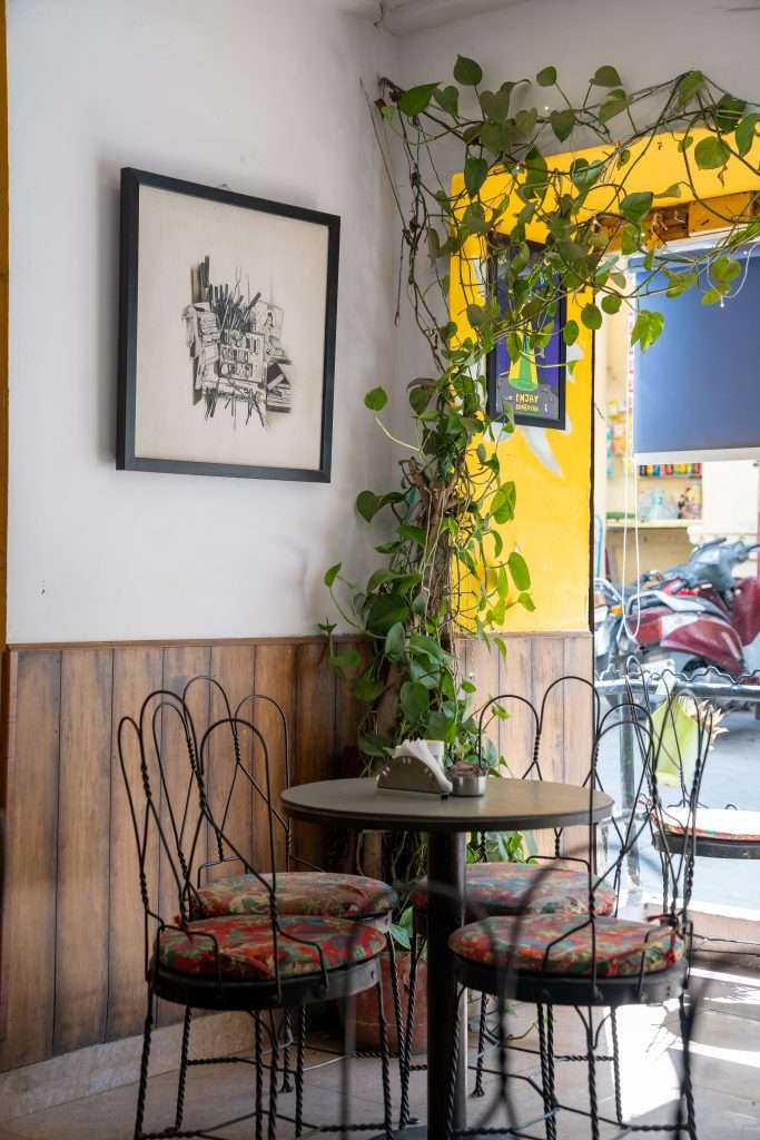 A cozy café corner featuring a round table and two metal chairs with colorful cushions. A potted plant with trailing vines decorates the space. Artwork hangs on the white wall above the table. Light streams in from a window with a view of parked motorcycles outside.Cafe Edelweiss Udaipur 