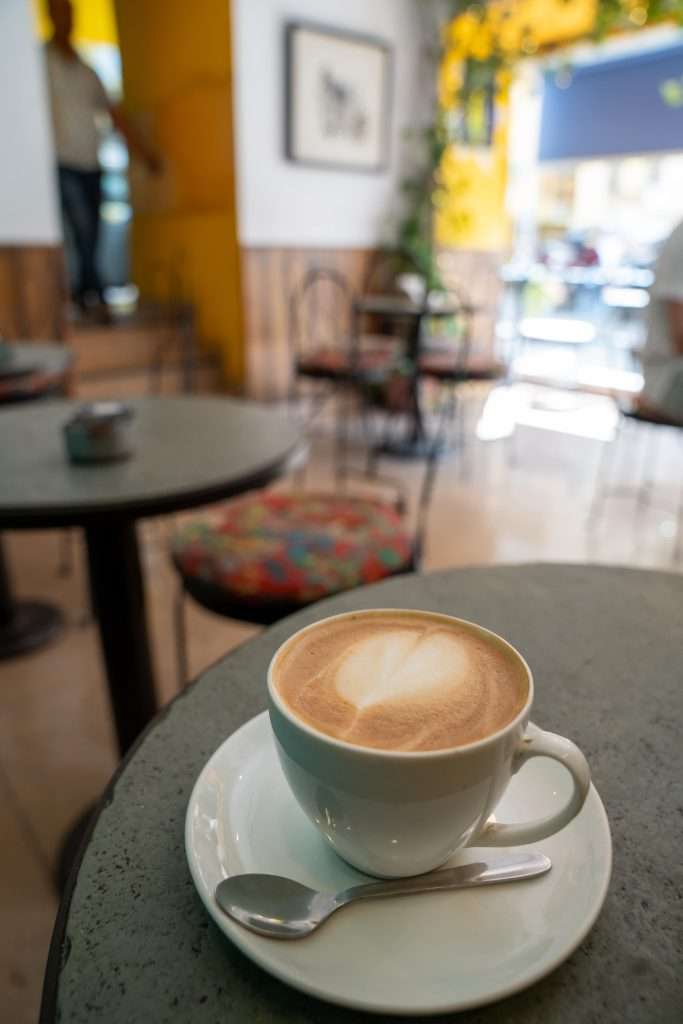 A close-up of a cup of coffee with intricate latte art on a saucer with a spoon, placed on a table in a cozy cafe. The background features blurred tables, chairs, and a person standing, creating a warm and inviting atmosphere. Cafe Edelweiss Udaipur 