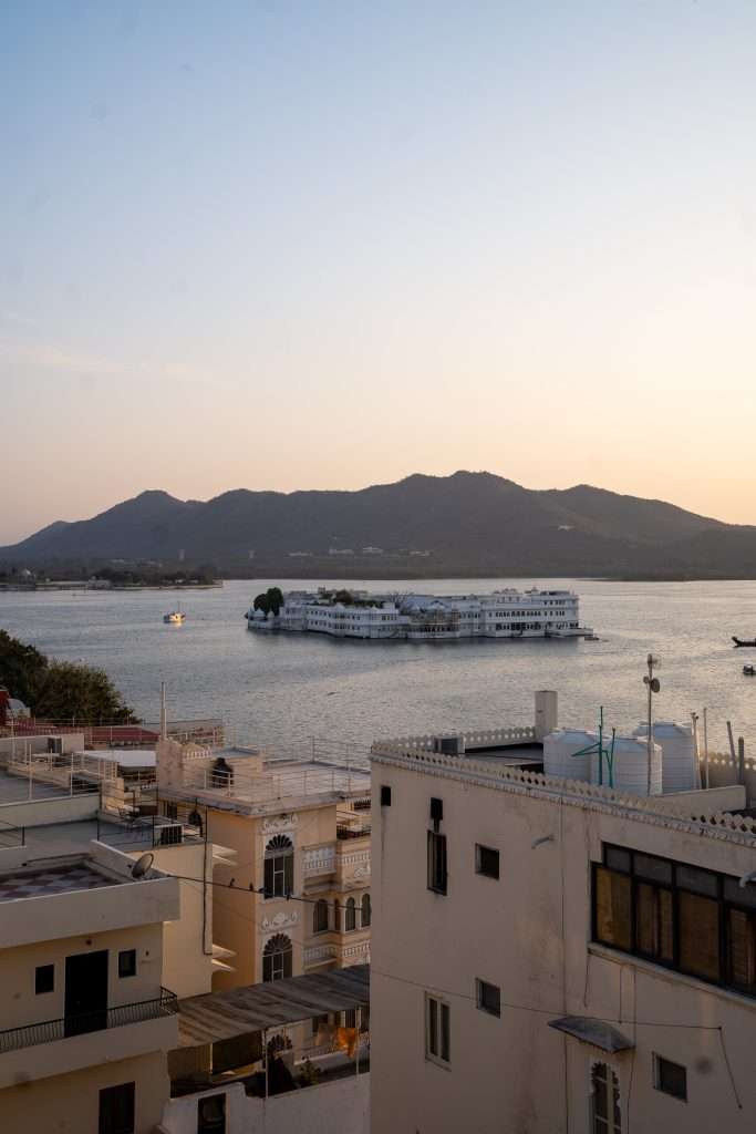 A serene view of a lake surrounded by mountains at sunset. A large, white building sits on the water with a boat nearby. In the foreground, there are buildings with intricate architectural designs and flat roofs. The sky is clear with a warm, golden glow - View of Lake Pichola in Udaipur