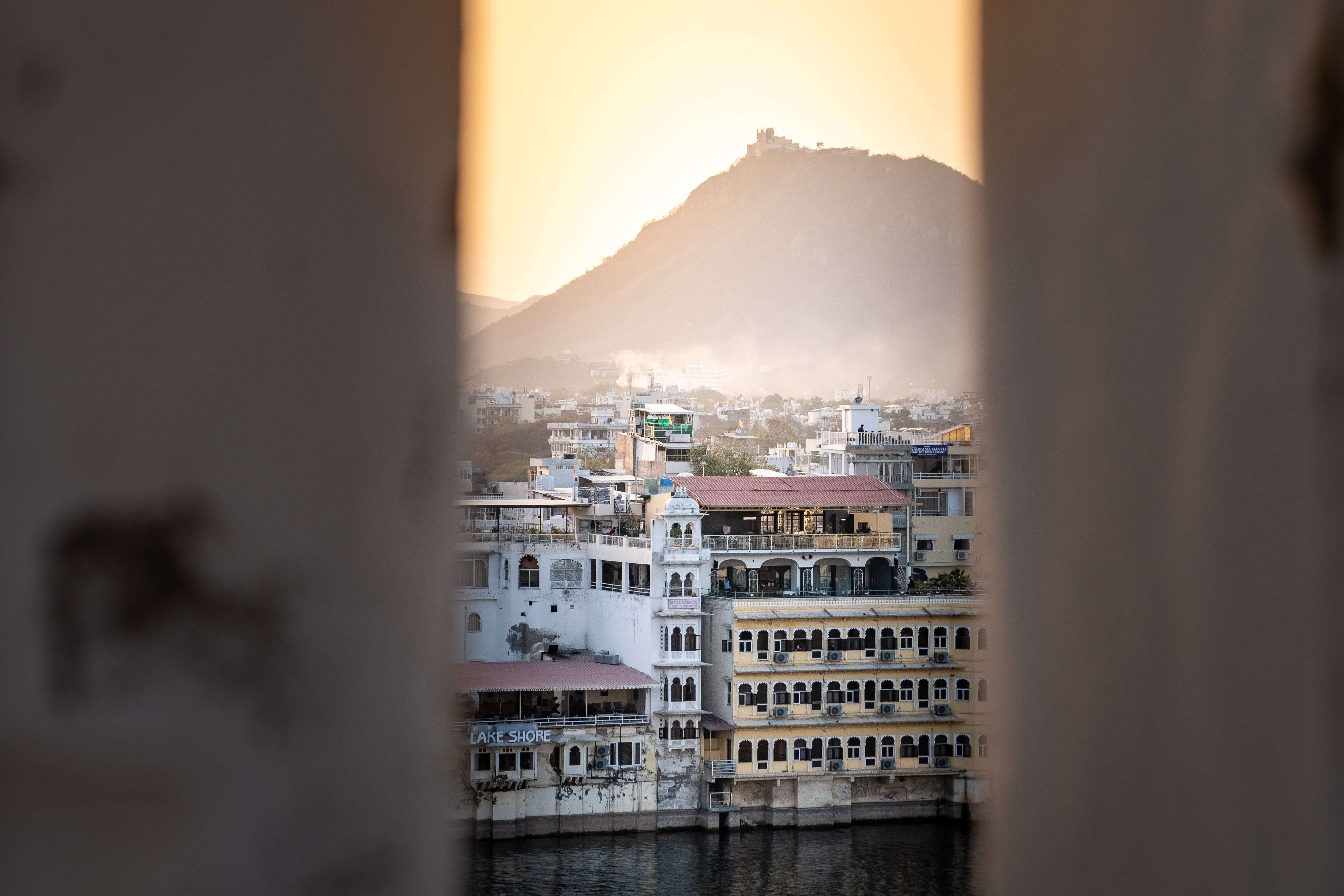 A picturesque view of a city with historic, multi-story buildings along a waterfront, seen through a narrow gap. The city is bathed in warm, golden light from the setting sun, with a hazy hill featuring a fort in the background—indeed making you wonder, is Udaipur worth visiting?