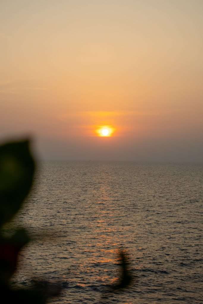 A serene sunset over a calm ocean, with the golden sun hovering just above the horizon, casting a warm reflection on the water. In the foreground, a blurred silhouette of a leaf is partially visible. The sky is a soft gradient of orange and yellow tones.