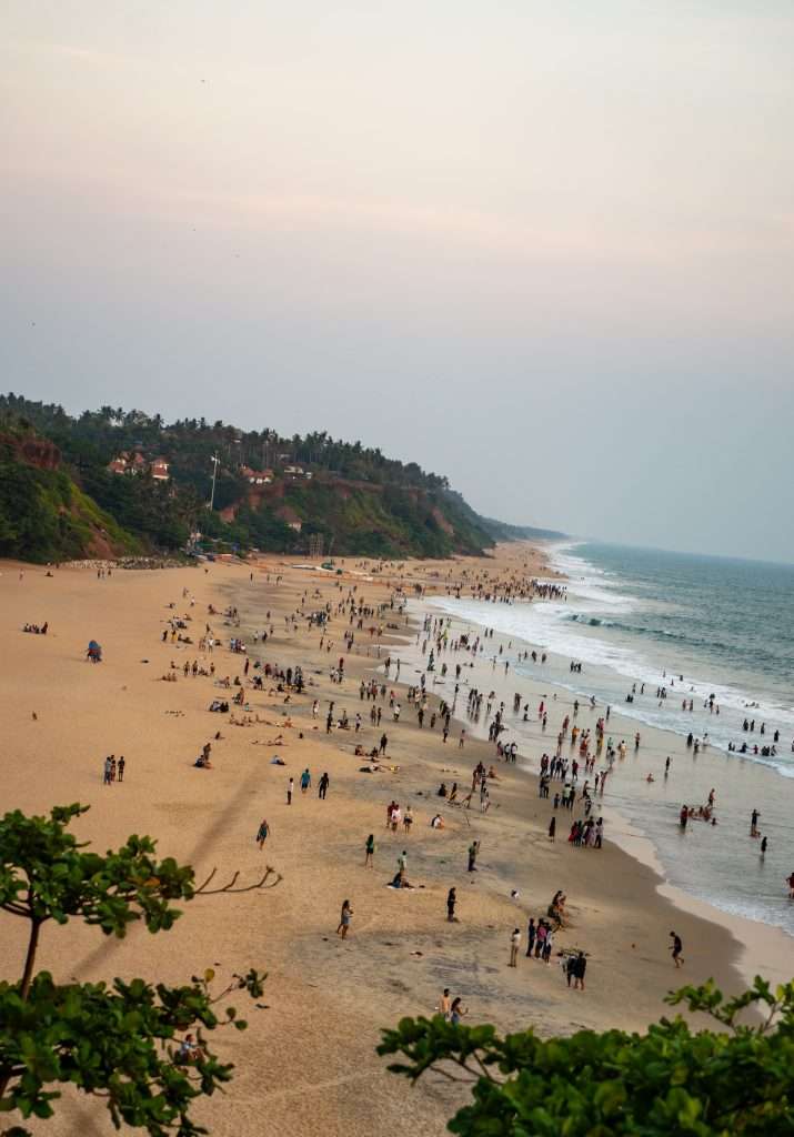 A bustling beach scene during sunset with numerous people walking, playing, and relaxing on the sand. The shoreline extends into the distance, bordered by waves on one side and greenery on the other, with buildings visible atop a hill.