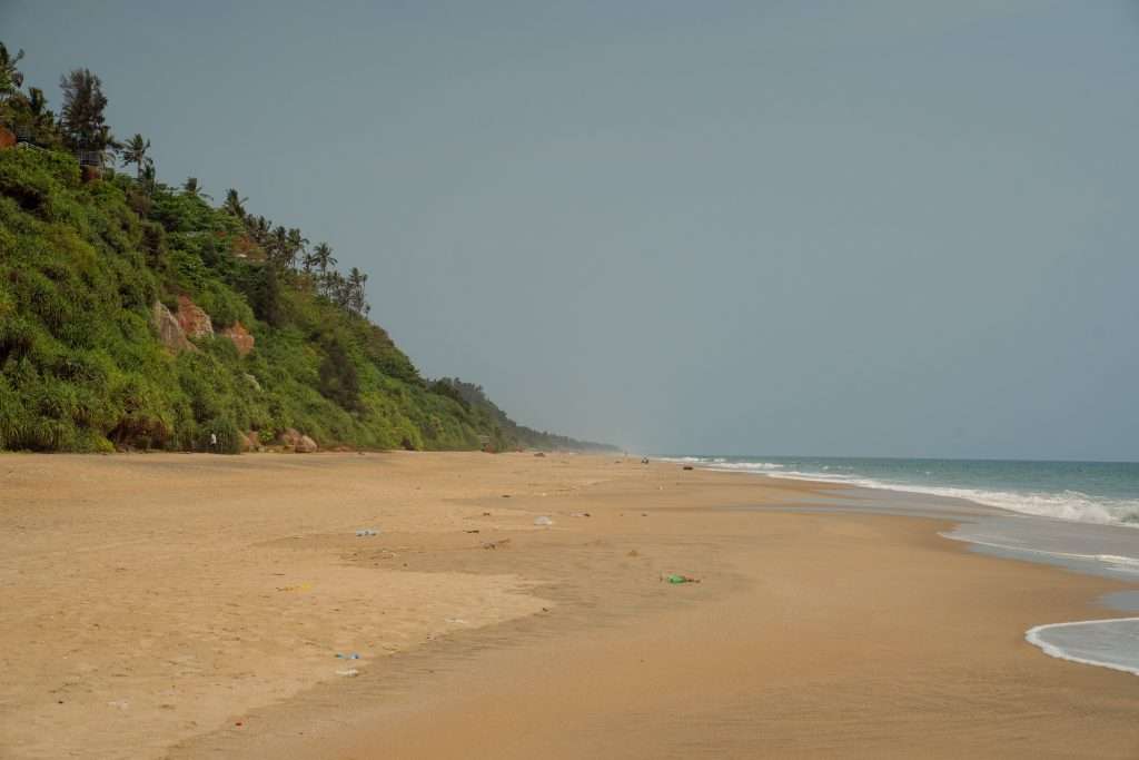 A serene, sandy beach with gentle waves lapping against the shore. Dense green foliage grows on a hillside to the left, extending down towards the beach. The sky is clear, creating a peaceful atmosphere with no people in sight.