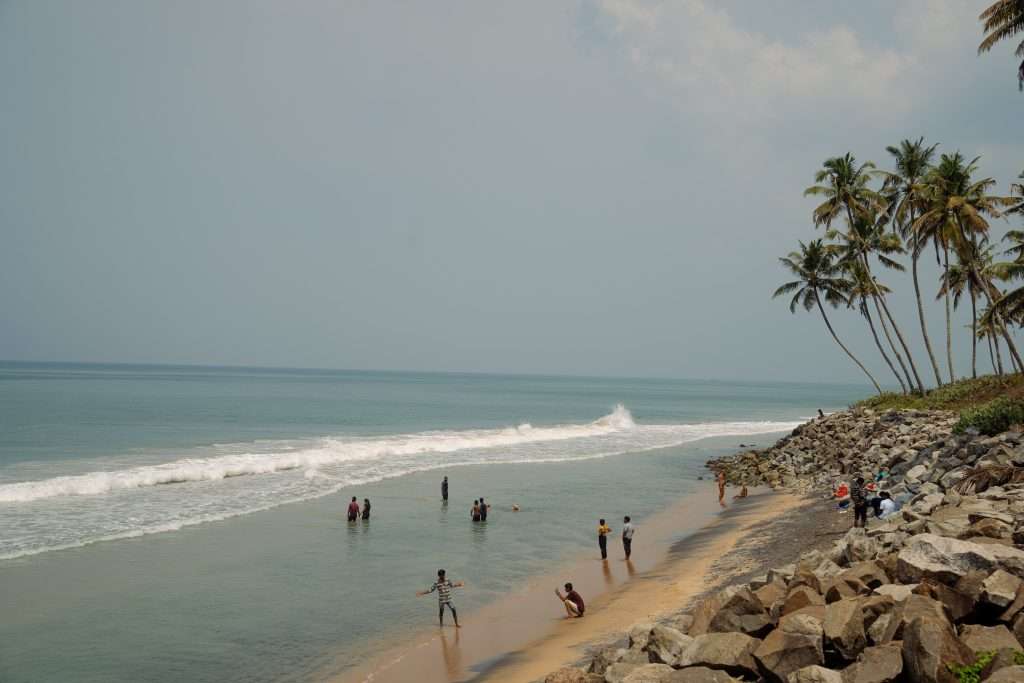 A sandy beach with scattered people enjoying the water. Some are swimming while others are standing on the shore. The sky is hazy, and palm trees can be seen on the right side along a rocky embankment. Waves roll gently towards the shoreline.