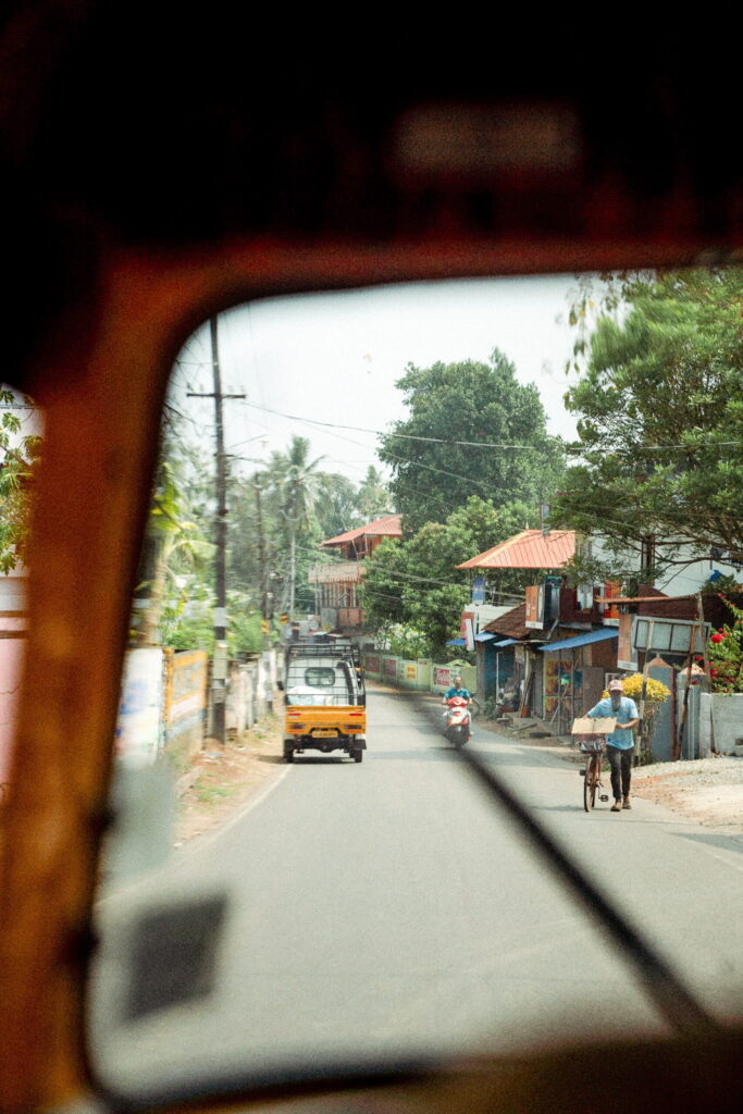 View from inside an auto rickshaw on a tranquil street lined with trees and houses, ideal for a 1-week Kerala itinerary; outside, a small truck, motorbike, and man pushing a bicycle add to the local charm.