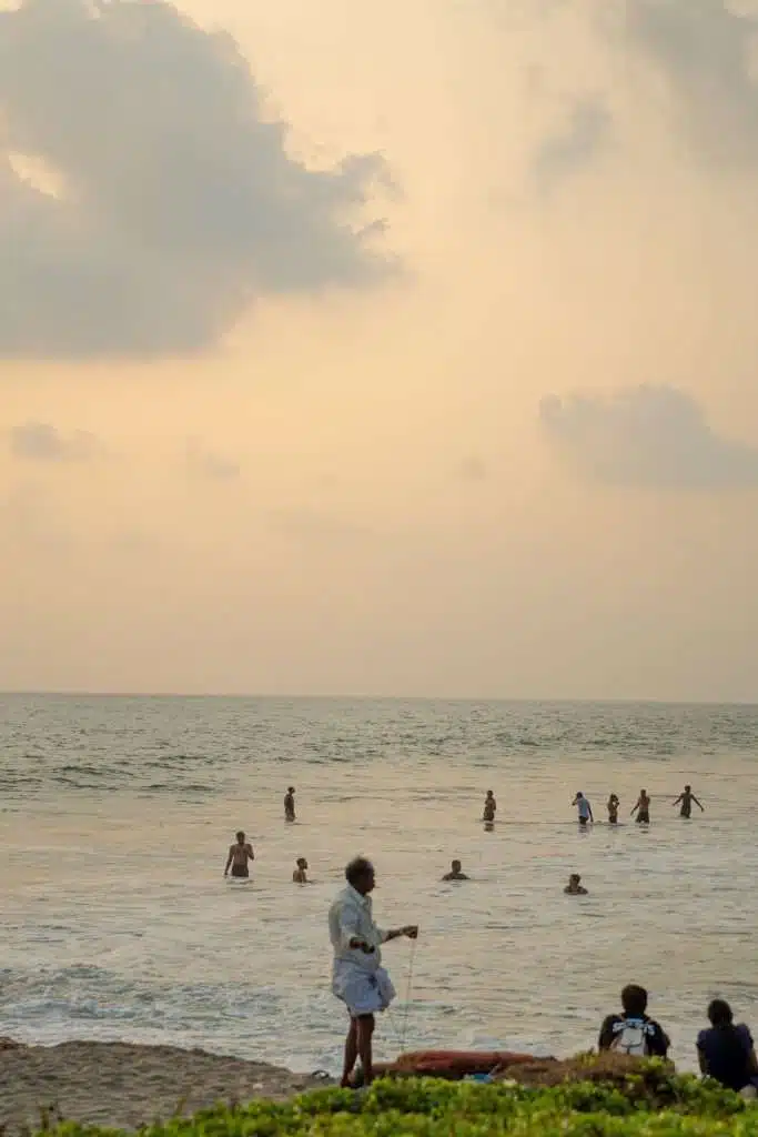 People enjoy a beach scene during sunset, with some swimming in the ocean and others relaxing on the shore. The sky is filled with soft clouds, creating a tranquil atmosphere. A person stands in the foreground while others sit nearby. Greenery borders the bottom of the image.