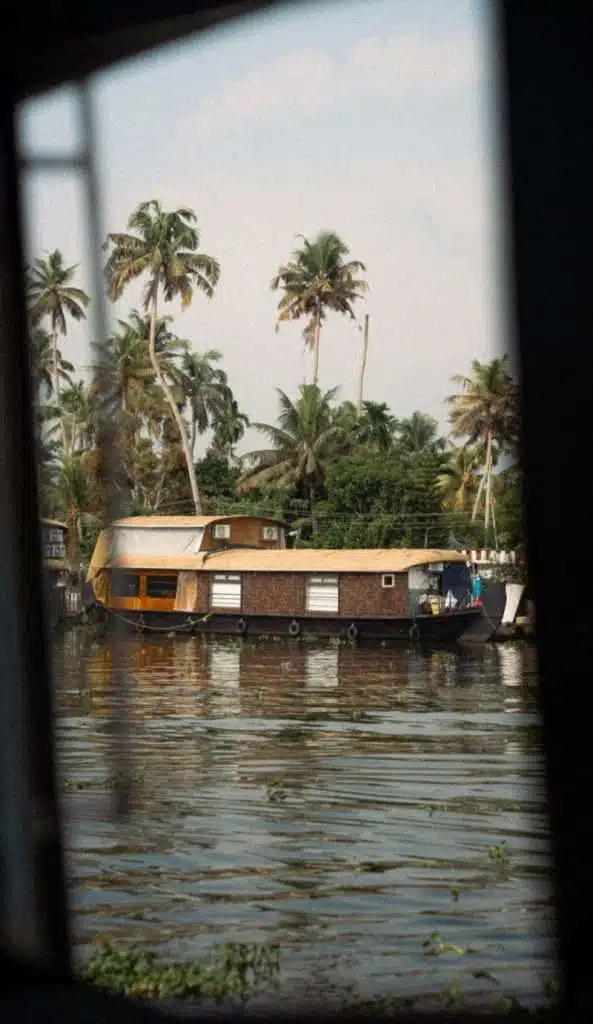 View of a serene water body framed by an open window, showcasing a traditional houseboat with a thatched roof and large windows, surrounded by tall palm trees and greenery. The scene reflects a calm, tropical atmosphere. Alleppey Backwaters Kerala. 