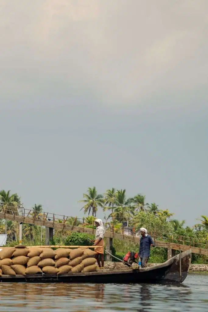 Men stand on a wooden boat loaded with sacks on a calm body of water, with lush green palm trees and a small wooden bridge in the background against a cloudy sky.