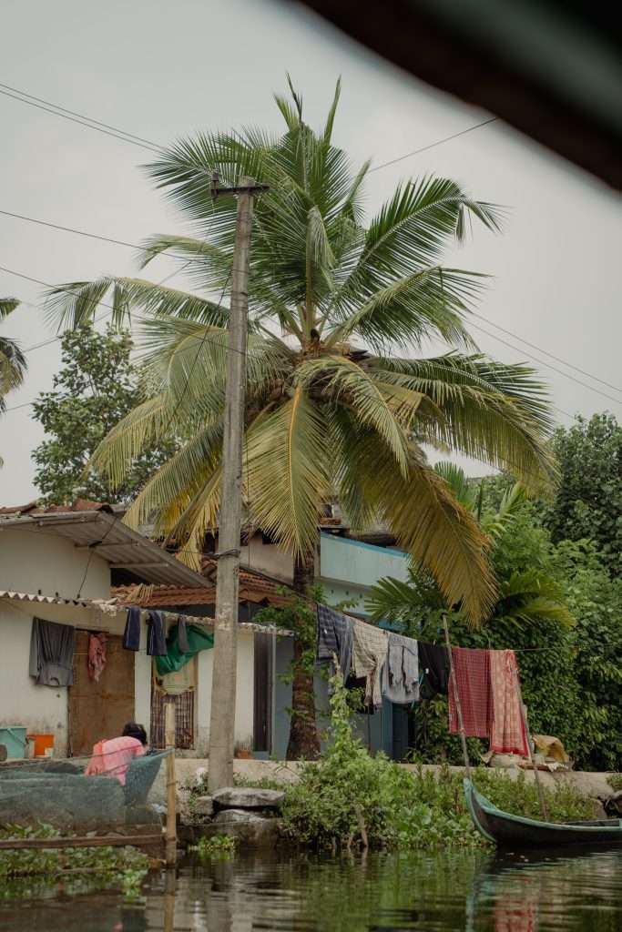 A small, rustic house with a palm tree in front, laundry hanging on a line, and a tranquil, narrow waterway reflecting the scene. A moored canoe is partially visible, and greenery surrounds the area, creating a serene rural setting.