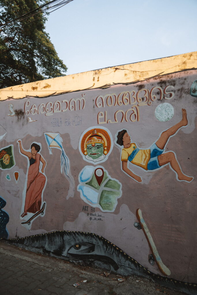 A colorful mural painted on a wall features a woman in a sari, a boy flying with a kite, a Kathakali dancer’s face, and "Kerala for High"—capturing the vibrant spirit of any 1-week Kerala itinerary.