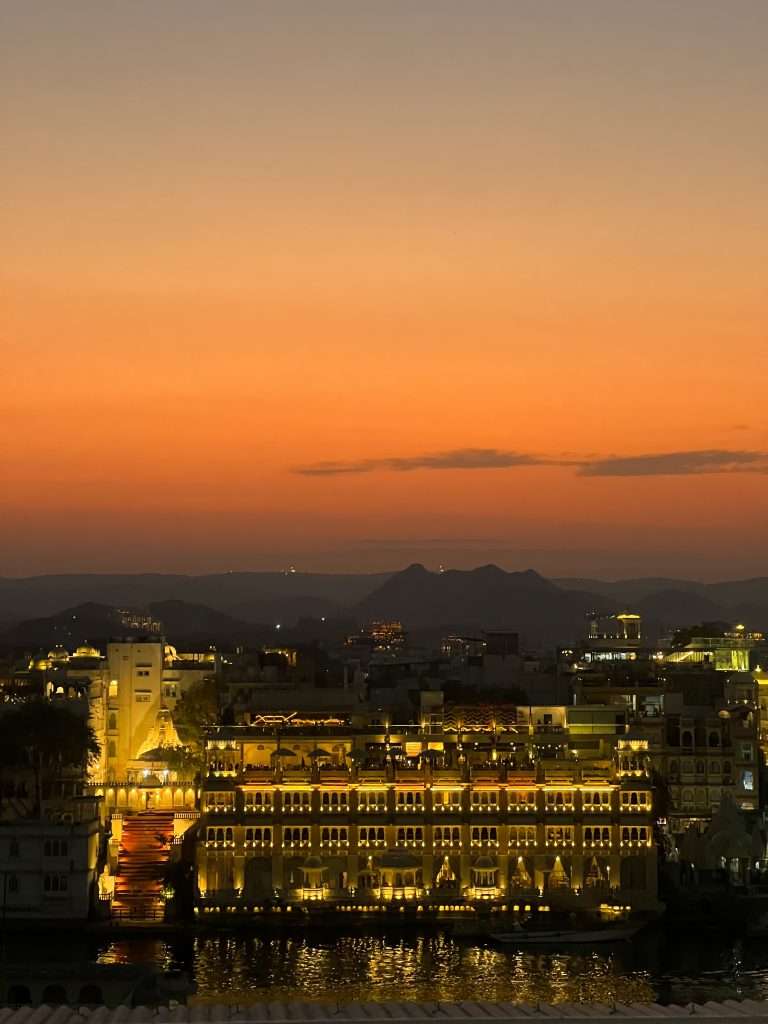 A beautiful sunset over a cityscape looking out over Udaipur featuring ornate buildings lit up with golden lights. The sky transitions from deep orange near the horizon to a lighter tone higher up, creating a stunning backdrop. The scene reflects in calm water in the foreground.