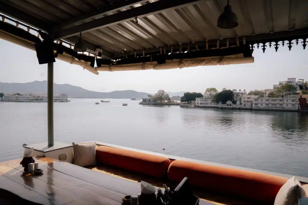 A covered outdoor seating area with a wooden table and red cushions overlooks a calm lake in Udaipur, where boats, trees, and distant buildings line the shore under a clear sky—showing exactly why Is Udaipur Worth Visiting is an easy question to answer.