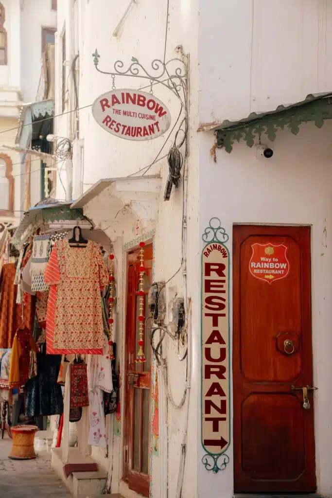 Narrow street with hanging colorful dresses for sale, a wooden door, and signs reading "Rainbow Restaurant" and "The Multi Cuisine Restaurant" on a whitewashed building—scenes like these make you wonder: Is Udaipur worth visiting? Absolutely.
