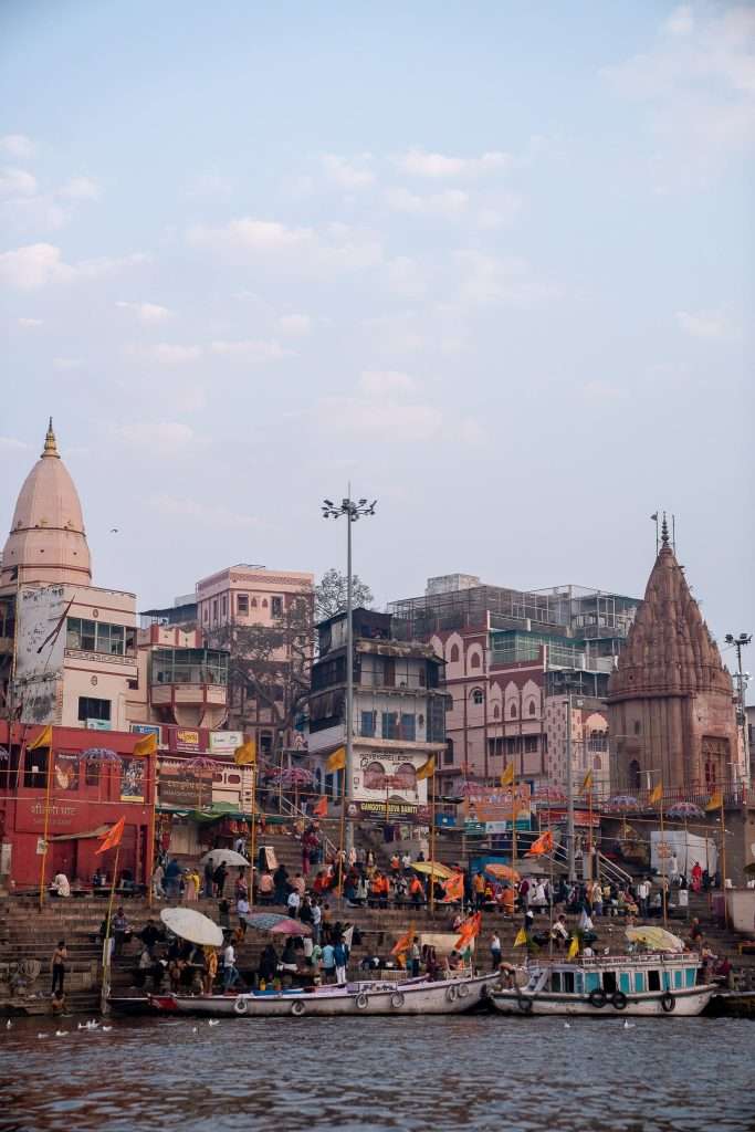 A busy riverside scene at a ghat in Varanasi, India, with people gathered on the steps and boats on the water—scenes like these answer the question: Is Varanasi worth visiting? The colorful buildings and temples under a partly cloudy sky say yes.