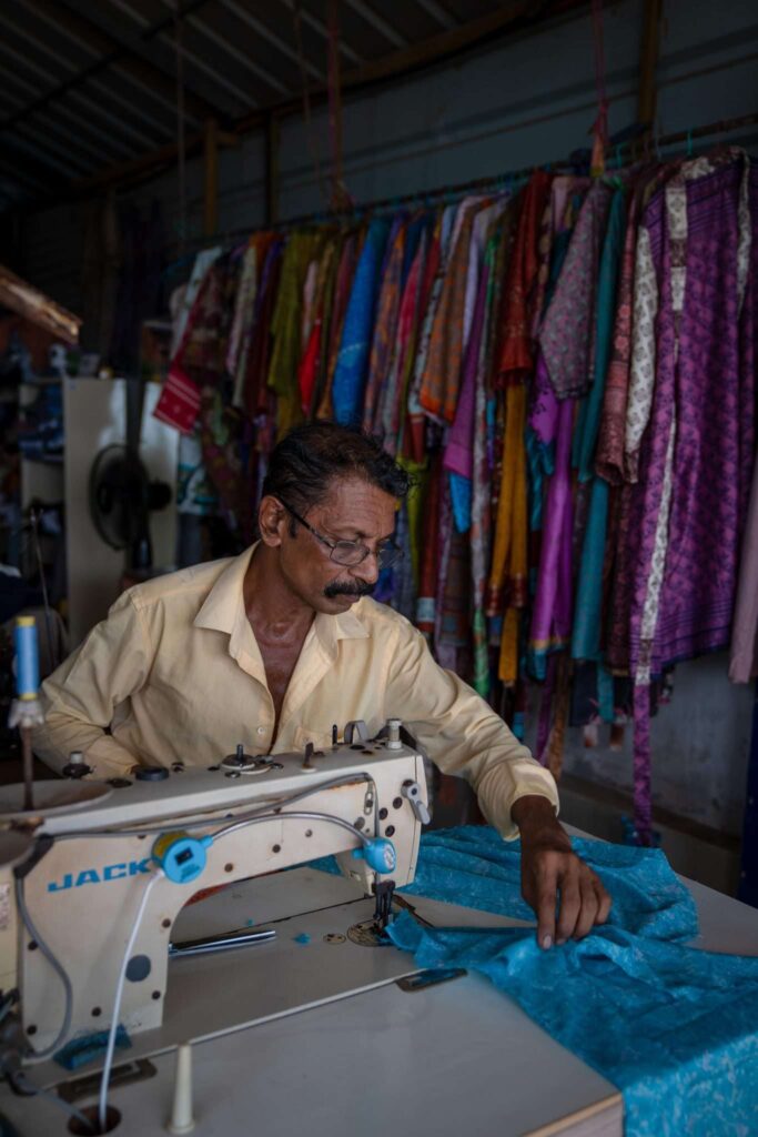 A man with glasses and a mustache sews blue fabric using a sewing machine in a shop, surrounded by colorful garments—like those you might find on a 1-week Kerala itinerary—hanging on racks behind him.