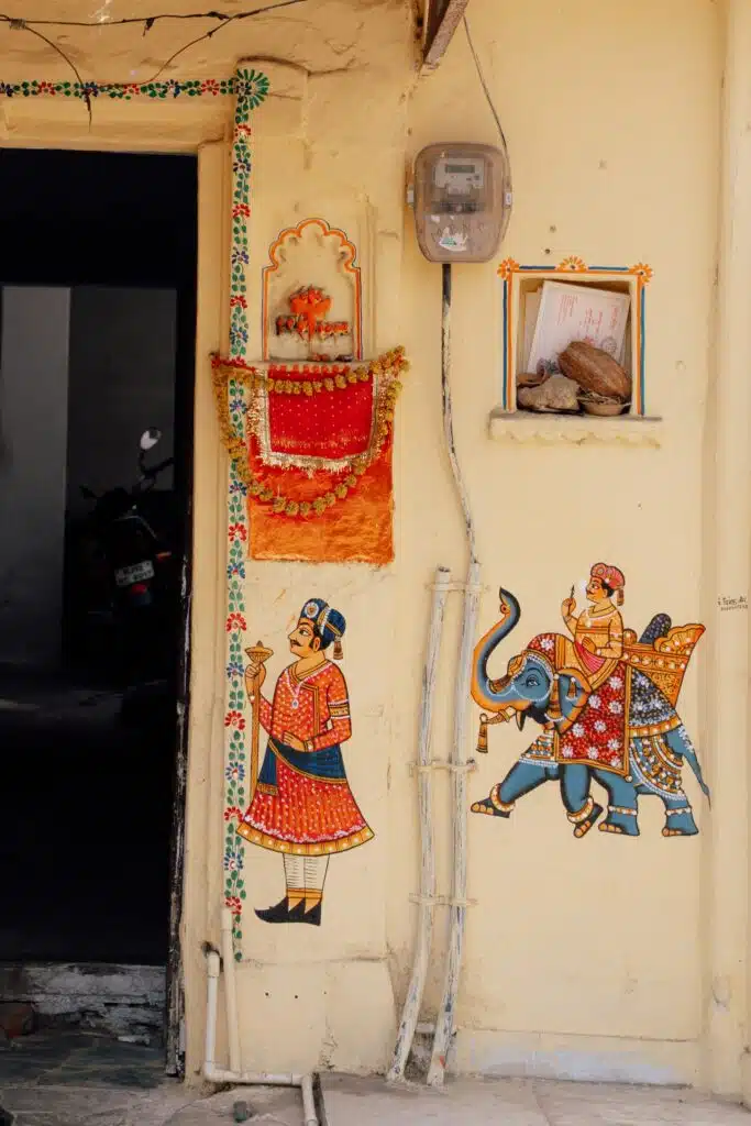 A yellow wall with colorful traditional Indian paintings—like a man with a staff and another on an elephant—garlands, a small ledge, and an electric meter above. Through the doorway, a parked motorcycle hints at why Is Udaipur Worth Visiting.