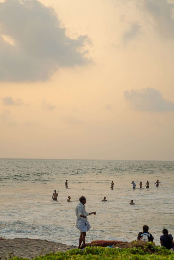 People swim and play in the ocean at sunset under a cloudy sky—a perfect scene for your 1-week Kerala itinerary. In the foreground, someone stands on the beach holding a fishing rod, while a few others sit nearby on the sand.