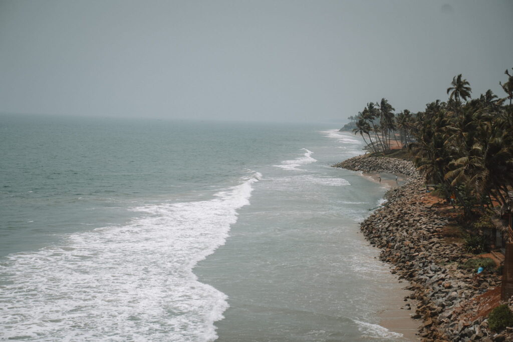 Waves crash onto a rocky shoreline lined with tall palm trees under a hazy sky, the coastline stretching into the distance—an idyllic scene perfect for any 1-week Kerala itinerary.