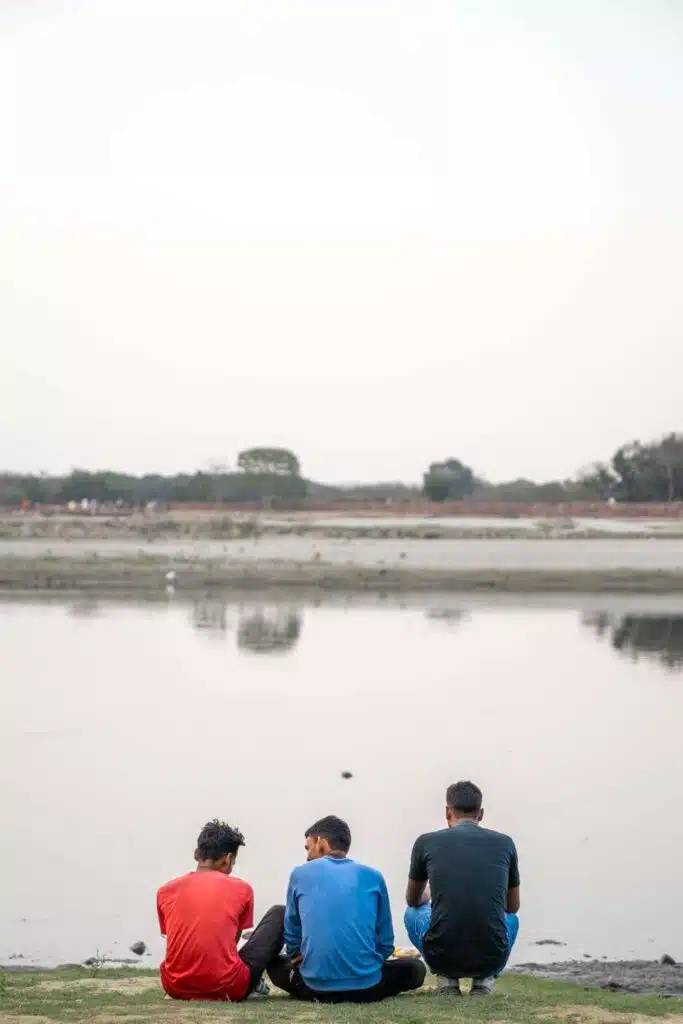 Three young men sit on the grass facing a calm river, with their backs to the camera, under a clear sky—perfectly capturing a tranquil moment along a 2-Week North India Itinerary. Trees and distant land are visible across the water.