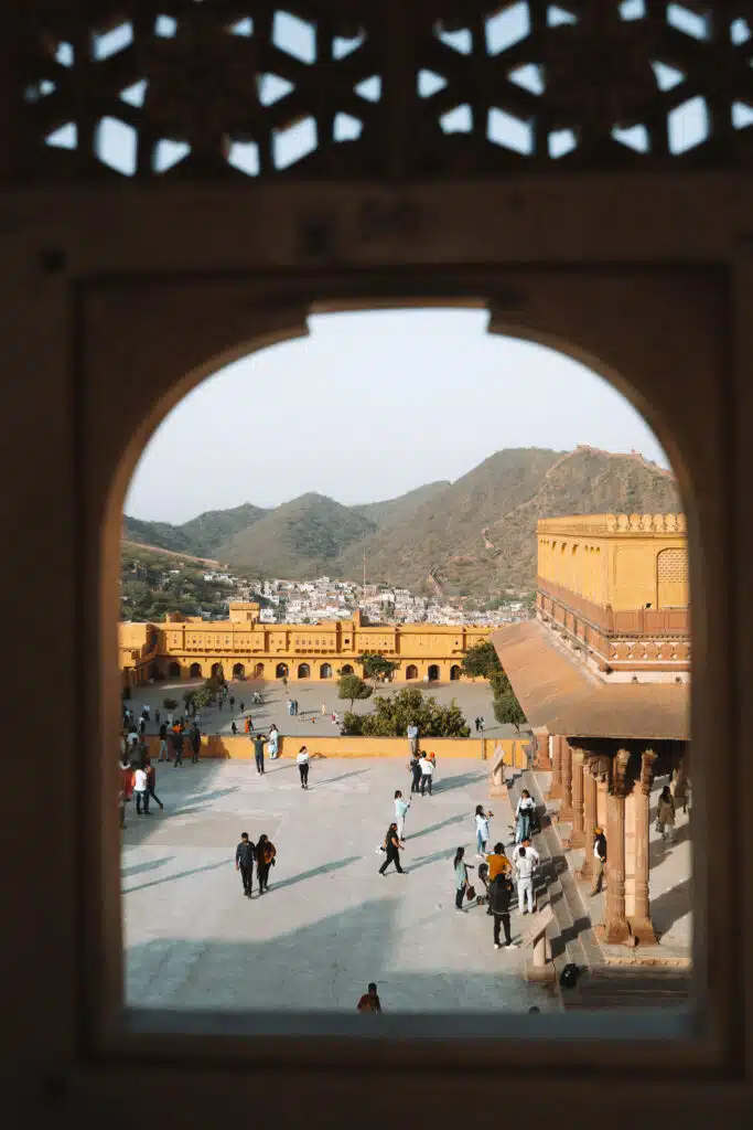 A view through an arched window shows people walking in a spacious courtyard of a yellow historic fort—an iconic stop on any 2-Week North India Itinerary—with mountains and a town in the background under a clear sky.