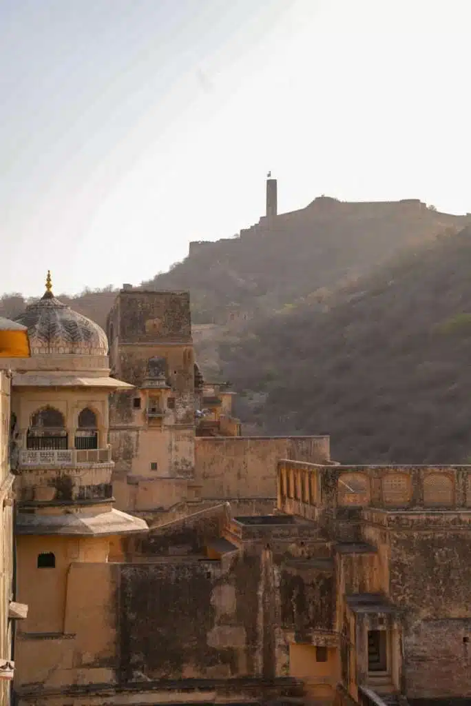 View of an old fort complex with domed and arched architecture in the foreground, perfect for a 2-Week North India Itinerary, set against a hillside with a large fortification and watchtower under a hazy sky.
