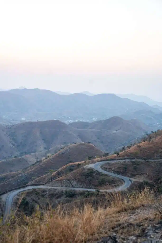 A winding road curves through dry, rolling hills with sparse vegetation—an inviting scene that could be part of your 2-Week North India Itinerary. Mountains stretch into the distance beneath a pale, hazy sky at dusk or dawn.