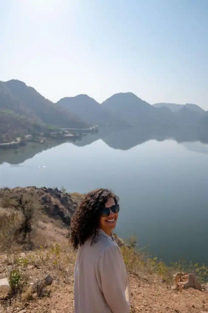 A woman with curly hair and sunglasses smiles while standing on a rocky cliff, capturing the spirit of adventure—a perfect moment for inspiration on a 2-Week North India Itinerary exploring tranquil lakes and distant mountains.