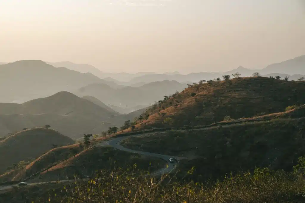 A winding road curves through rolling hills covered in sparse vegetation, evoking the landscapes found on a 2-Week North India Itinerary, with misty mountains fading into the distance under a soft, pale sky at sunrise or sunset.