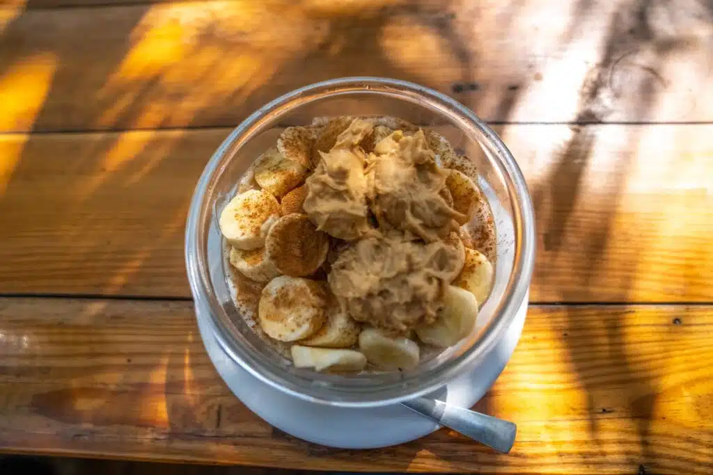 A glass bowl filled with sliced bananas, sprinkled with cinnamon, and topped with peanut butter sits on a white saucer beside a spoon, bathed in sunlight—an energizing snack perfect before planning your 2-Week North India Itinerary.