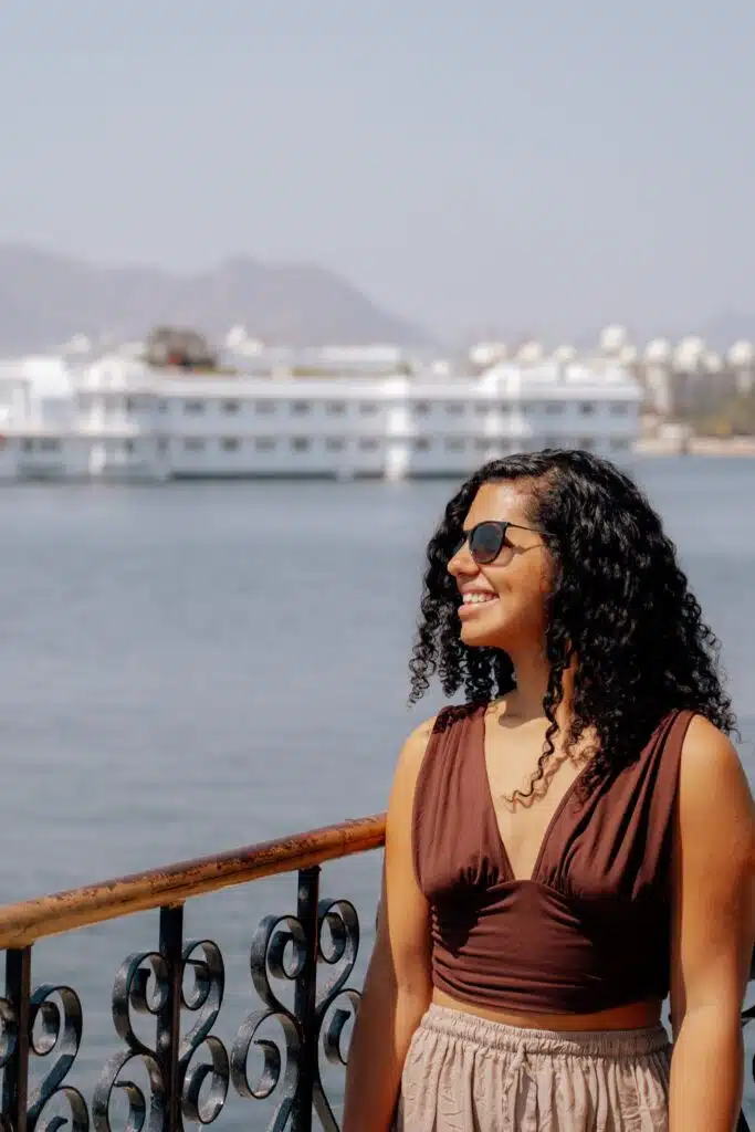 A woman with curly hair and sunglasses smiles while standing by a railing overlooking a lake—perhaps a dreamy stop on her 2-Week North India Itinerary—with a large white building and distant mountains in the background.