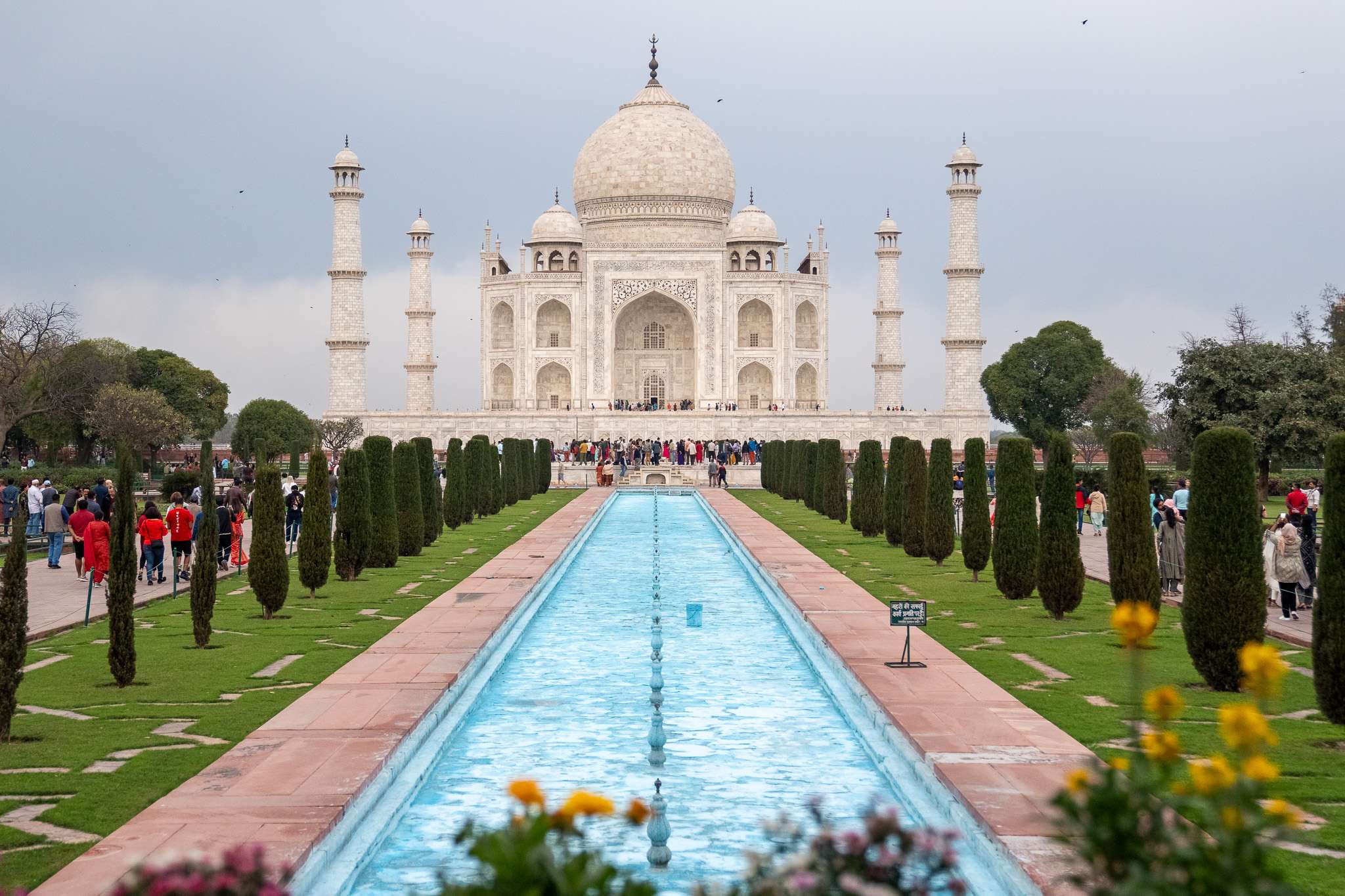 The image captures the Taj Mahal, an iconic white marble mausoleum featured in many 2-Week North India Itineraries. Its large dome and four minarets rise majestically, with a long reflecting pool stretching from the foreground, flanked by manicured gardens where visitors gather near the entrance.