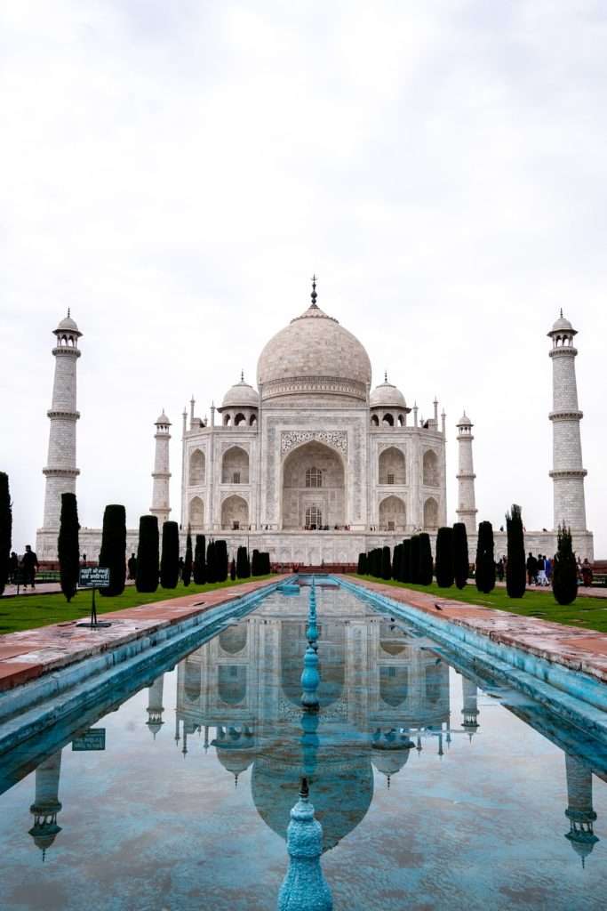 The image showcases the Taj Mahal, a white marble mausoleum with reflecting pools and symmetrical minarets, set against a cloudy sky. This iconic site is a highlight of any 2-week North India itinerary, emphasizing its architectural beauty and the serenity of its surrounding gardens.