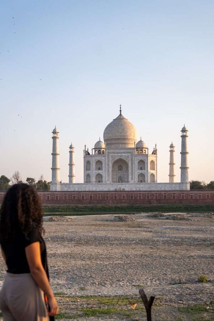 A person with long curly hair stands in the foreground, gazing at the Taj Mahal in the distance from Mehtab Bagh. Part of a 2-Week North India Itinerary, this moment unfolds as the sun sets, casting a warm glow on the iconic white marble mausoleum. The foreground is a dry, grassy area.