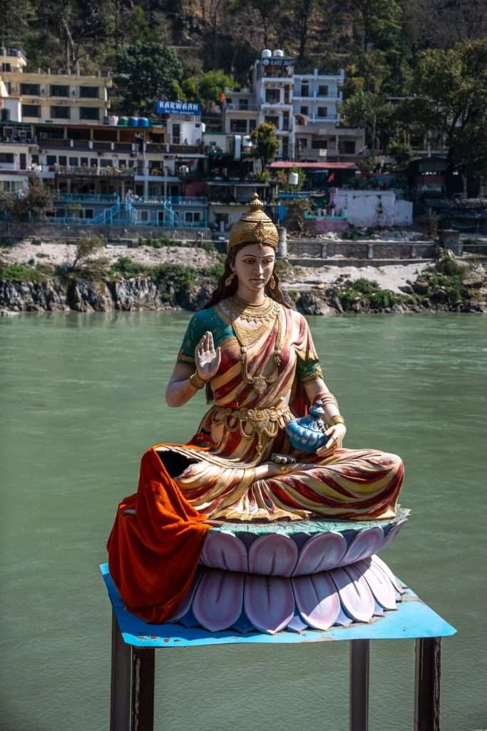 A statue of a seated deity with a serene expression and a raised hand gesture is perched on a lotus pedestal near a calm river. This picturesque scene, often featured in 2-Week North India Itineraries, is framed by hillside buildings and trees in the background.