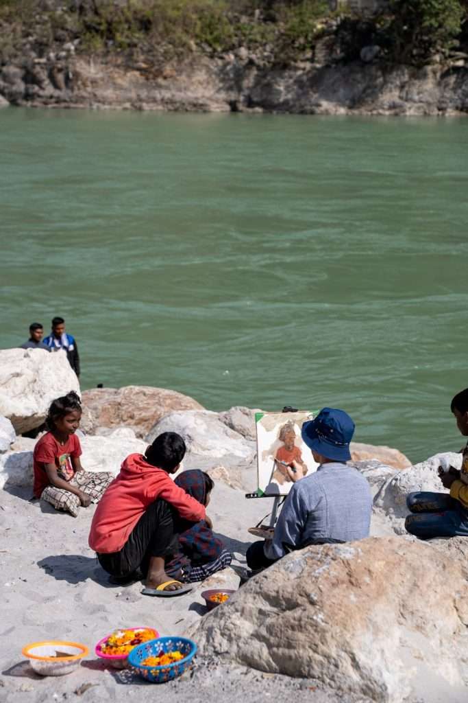 A group of people sits by a riverbank on rocks, with someone painting an artwork on an easel, capturing the serene beauty of their 2-week North India itinerary. Bright bowls with colored beads are nearby, while the river flows gently in the background as a few stand by the water's edge.