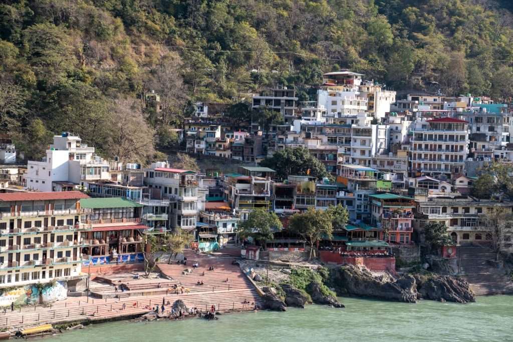 Experience a scenic view reminiscent of a 2-Week North India Itinerary, with buildings nestled on a hillside beside a river. The colorful multi-story architecture harmonizes with stone steps leading to the riverbank, surrounded by lush greenery and a dense cluster of trees in Rishikesh India. 