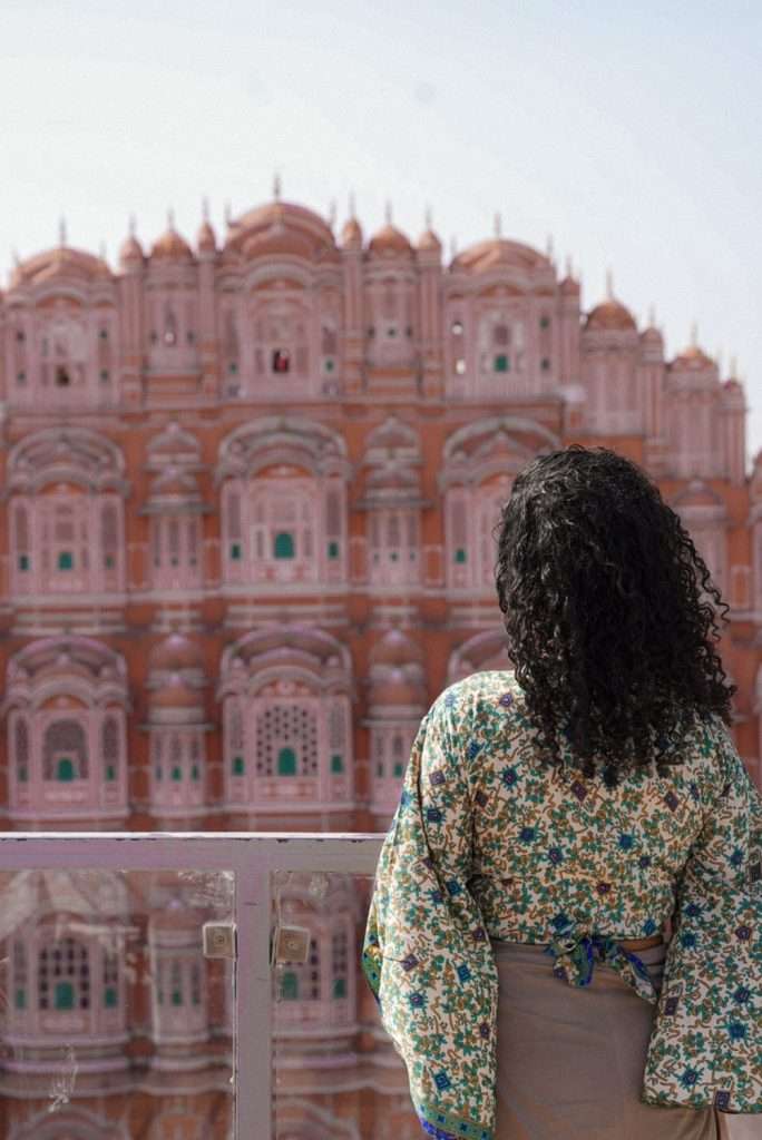 A person with curly hair and a floral top stands facing the majestic Hawa Mahal, an iconic stop on any 1-Week Rajasthan Itinerary, its ornate pink facade gleaming under the clear sky.