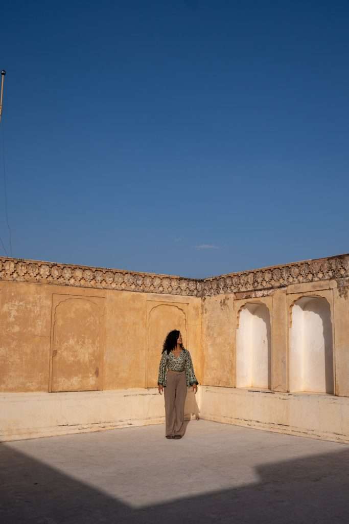 A person stands in a sunlit, open courtyard with high, ornately patterned walls under a clear blue sky, just like those featured in a 1-Week Rajasthan Itinerary. The walls are adorned with intricate designs, and the person gazes towards the corner of the space.