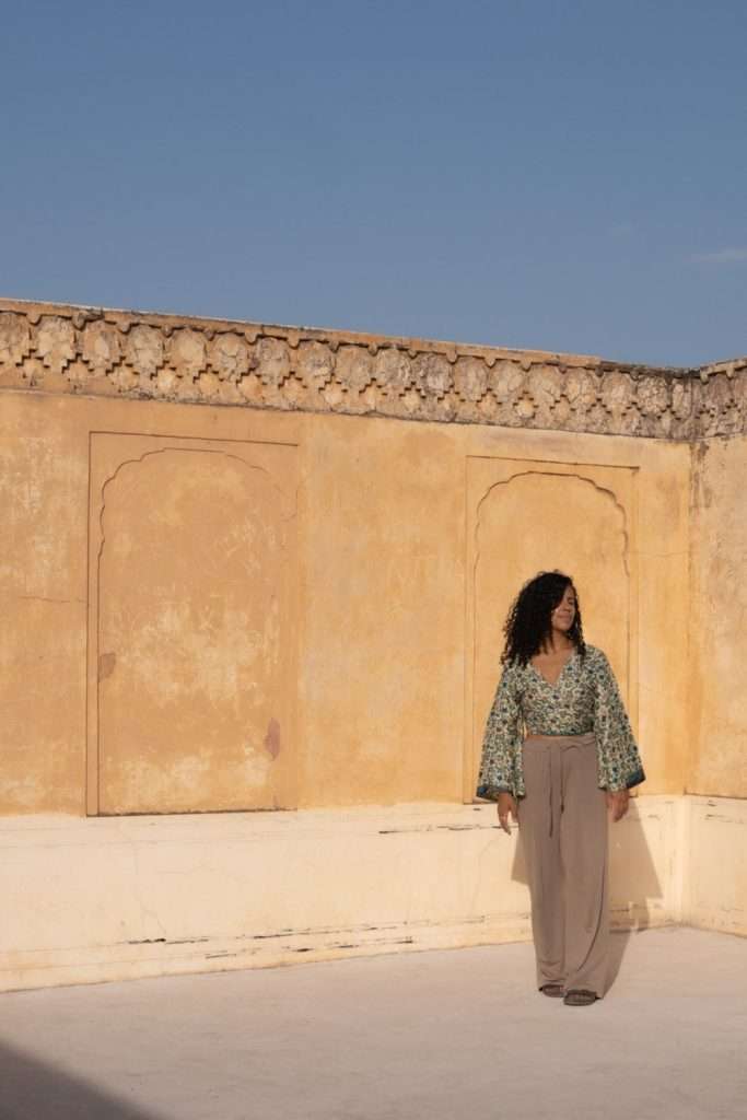 In a sunlit courtyard on their 1-week Rajasthan itinerary, a person with long curly hair stands against a rustic yellow wall adorned with ornate carvings. Dressed in a green patterned top and beige pants, they bask under the clear blue sky.