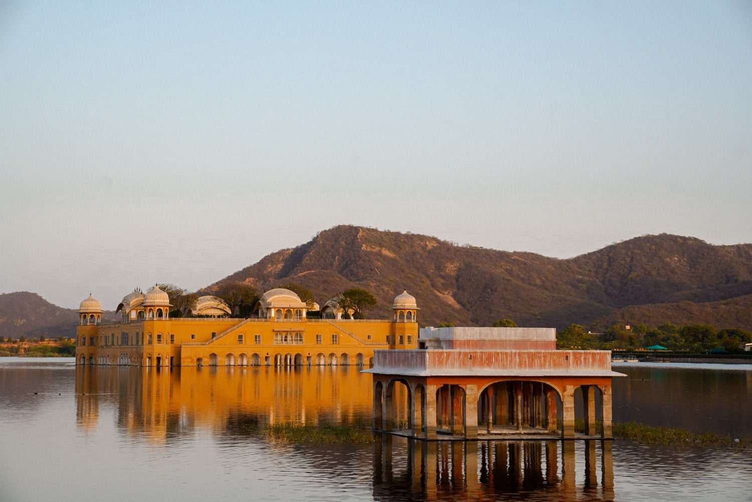 The image, a highlight of a 1-Week Rajasthan Itinerary, shows Jal Mahal—a yellow palace with domes—nestled amid a calm lake. Surrounding hills rise under a clear sky, reflecting the golden hue of the setting sun on the serene water's surface.