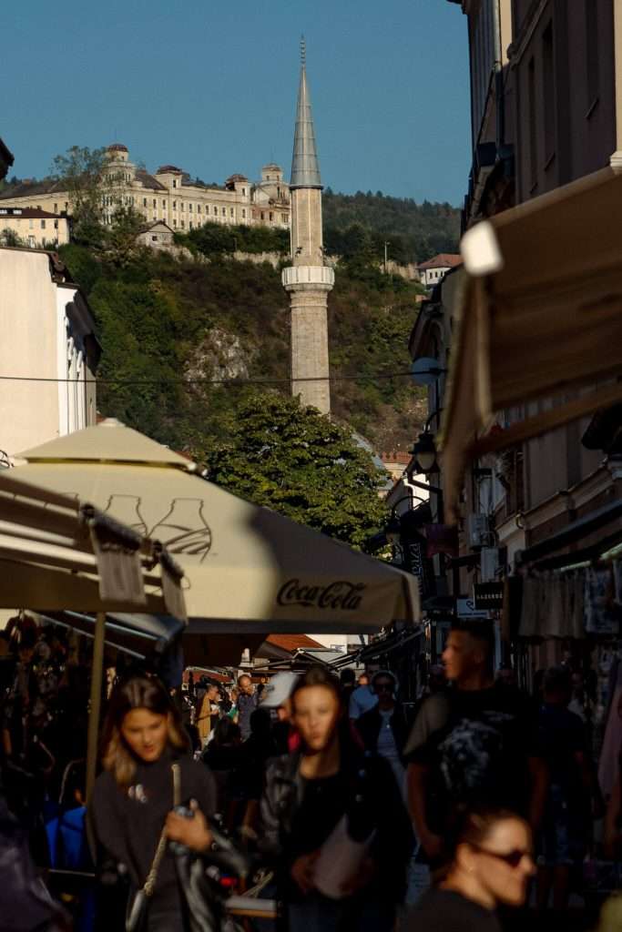 A bustling street scene unfolds in a lively one-day Sarajevo itinerary, with people walking under umbrellas. A mosque's tall minaret rises atop a hill in the background, surrounded by greenery and buildings. The atmosphere is vibrant and full of life.