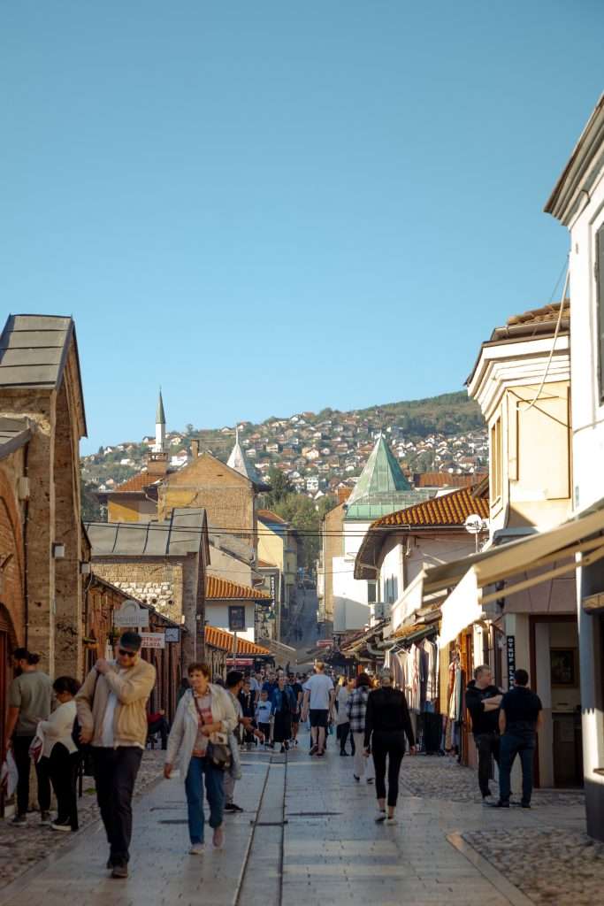 A bustling street scene in Baščaršija unfolds,. People stroll along the cobblestone pathway lined with shops. In the background, scenic hills and traditional architecture, including a prominent mosque with a tall minaret, stand under a clear blue sky.