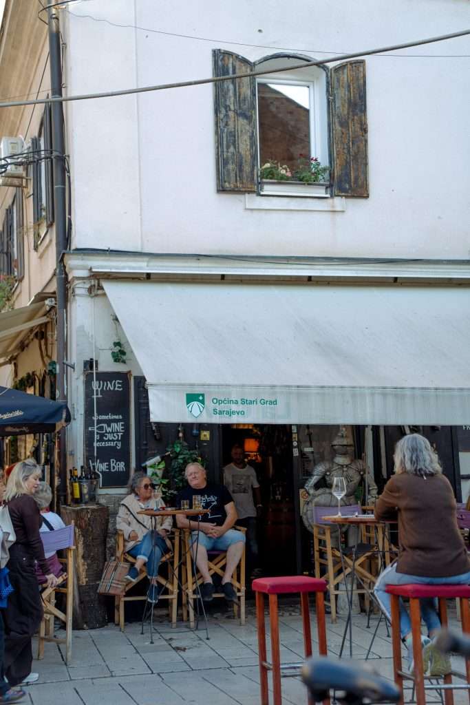 People enjoy a quaint cafe on a cobblestone street, perfect for a one-day in Sarajevo itinerary. The white building features wooden shutters and a chalkboard menu. A person stands in the entrance, while potted plants adorn the window ledge above tall tables and stools.