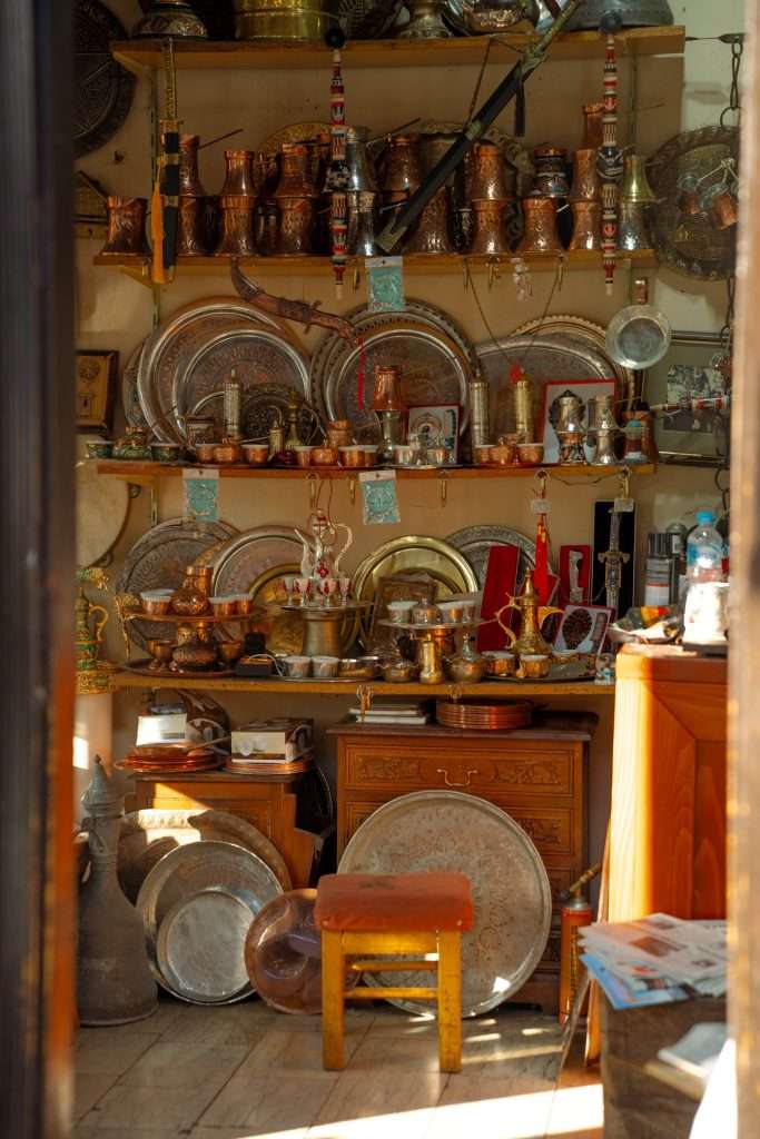 A cozy shop in your one-day Sarajevo itinerary is filled with decorative metalware, including plates, teapots, and lanterns on shelves. A small orange stool sits at the center, with sunlight streaming through the doorway, creating a warm and inviting atmosphere.