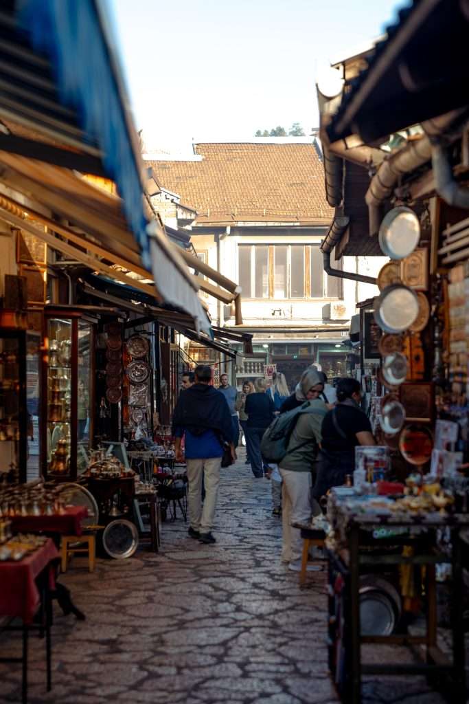 A narrow, cobblestone street in Sarajevo is lined with small shops displaying various goods, including pottery and trinkets. As part of a one-day itinerary, people stroll along, browsing the items under a clear sky with sunlight casting playful shadows on the ground.