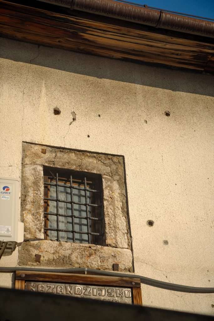 A small window with metal bars is set in an old, cream-colored wall marked by cracks and holes, echoing the charm of a one-day in Sarajevo itinerary. Below the window is a sign with a word, while part of an air conditioning unit peeks out on the left side.